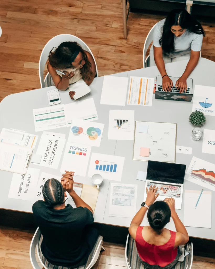Overhead view of three marketing professionals sitting around a table covered with laptops, printed charts, a marketing strategy document, trends report, and data sheets — representing a collaborative digital marketing planning session.