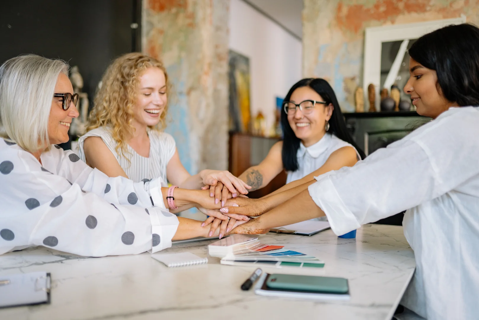 Four diverse female digital marketing professionals stacking hands together around a table with color swatches, symbolizing KMP Digital Media's team unity and genuine partnership as a digital marketing agency